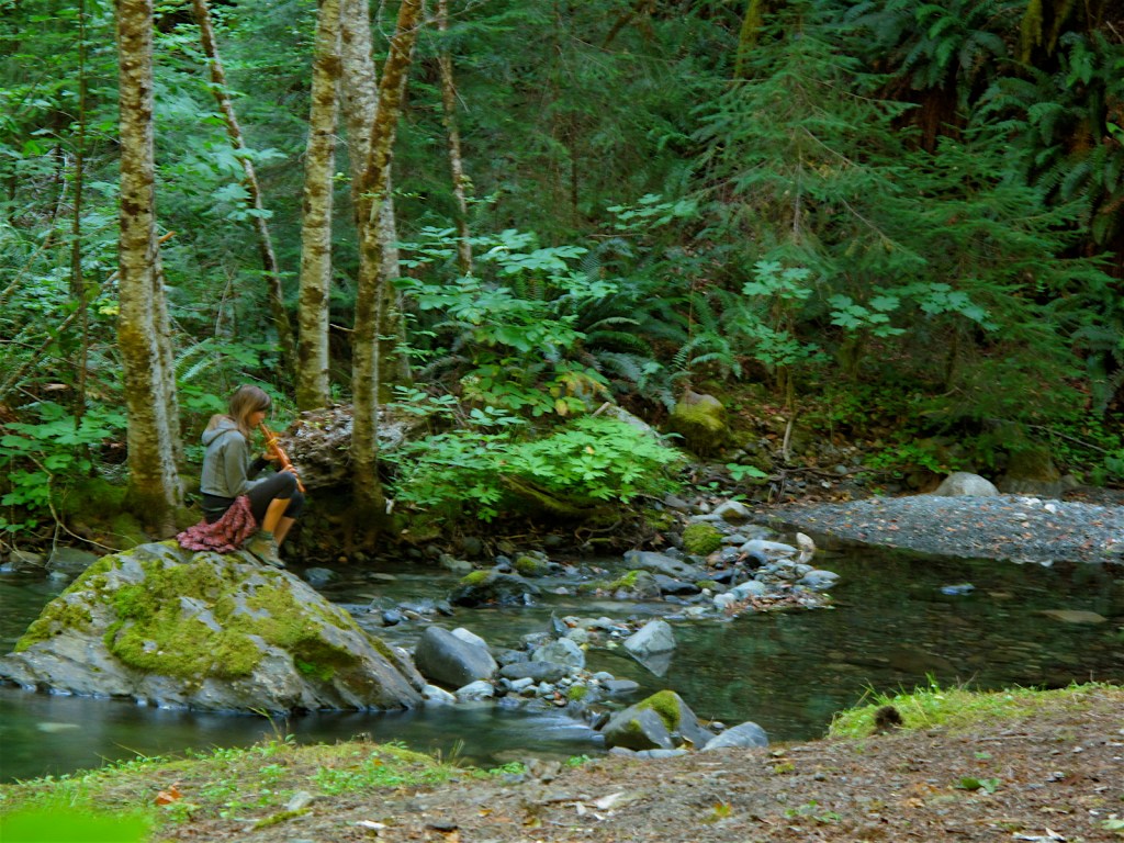 Jen Poole playing flute on a mossy rock beside a forest creek